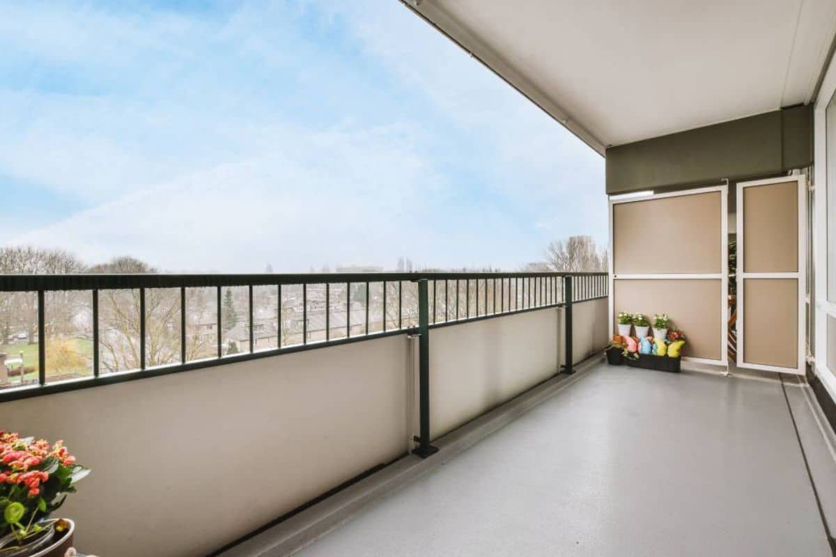 Spacious balcony with a clear view of trees and a blue sky. Potted flowers line the railing and corner, creating a serene and inviting atmosphere.