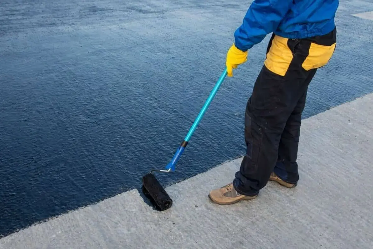 A person in protective clothing uses a blue-handled roller to apply black waterproof coating on a concrete surface, conveying focus and precision.